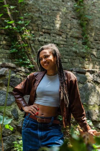 Happy african american woman in brown leather jacket standing with hand on hip - foto de stock