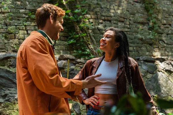 Cheerful man with red hair gesturing and talking with african american woman outside — Foto stock