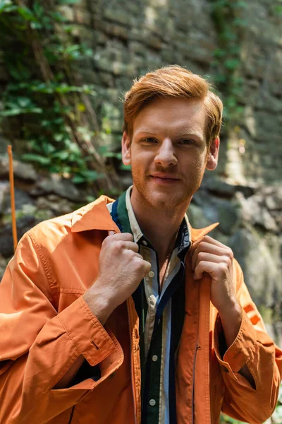 Joyful redhead man in orange jacket smiling and adjusting collar on jacket outside — стоковое фото