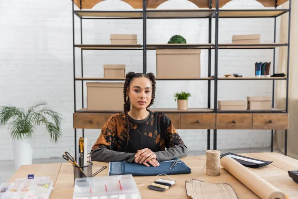 Young african american craftswoman looking at camera near cloth and sewing equipment in worktop — Stock Photo