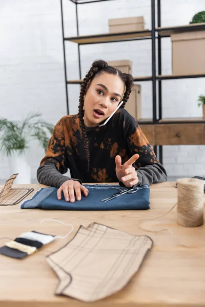 African american craftswoman talking on smartphone near cloth and sewing equipment in workshop — Stock Photo