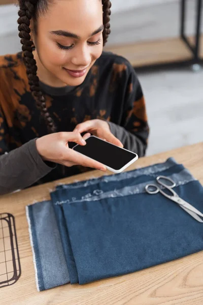 Smiling african american designer taking photo on smartphone near cloth and scissors in workshop — Foto stock