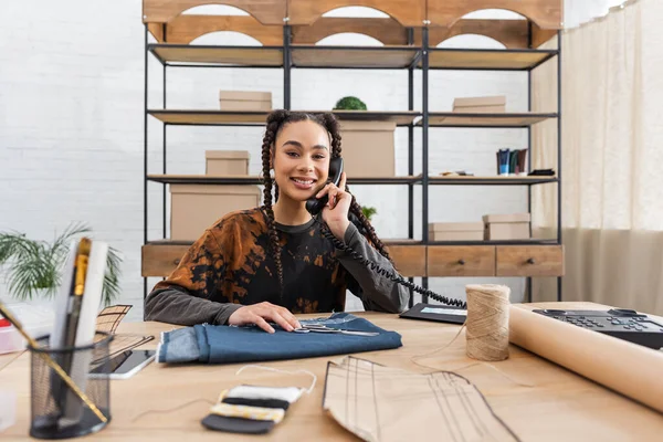 Smiling african american craftswoman talking on telephone near sewing equipment - foto de stock