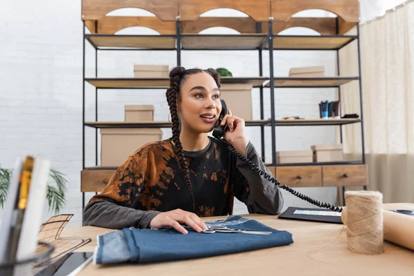 African american designer talking on telephone near cloth and scissors in workshop — Photo de stock