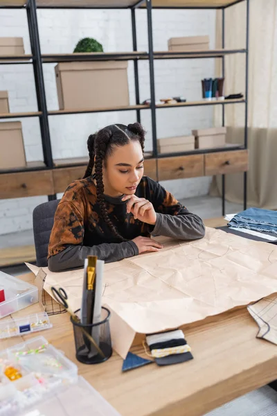 Focused african american craftswoman looking at sewing pattern near equipment in workshop — Stock Photo