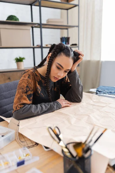 Pensive african american craftswoman looking at sewing pattern near equipment in workshop — Fotografia de Stock