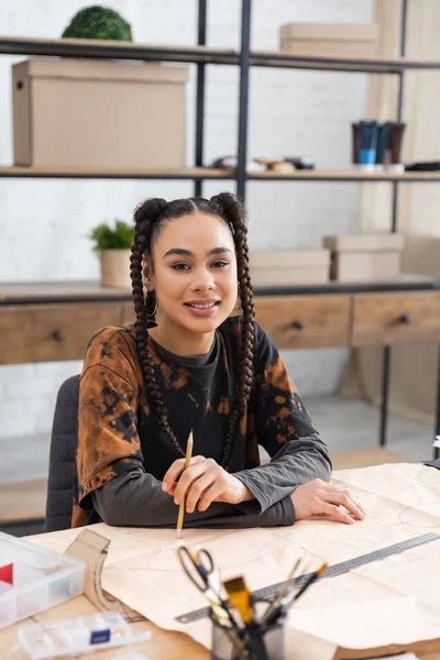 Positive african american craftswoman holding pencil near sewing pattern in workshop — Stock Photo