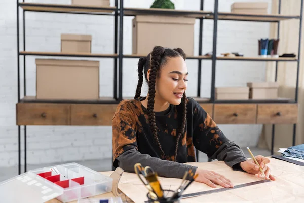 Smiling african american craftswoman drawing on sewing pattern near equipment — Stock Photo