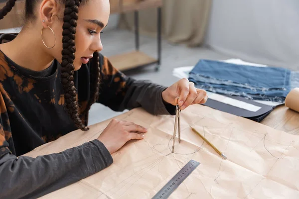 African american designer drawing with compass on sewing print near blurred cloth in workshop — Stock Photo