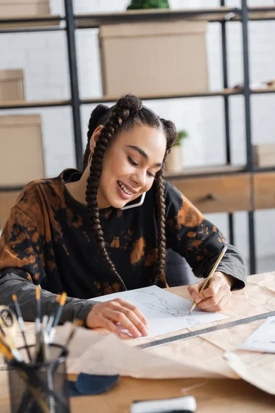 Smiling african american designer talking on smartphone and drawing sketch in workshop — Stock Photo