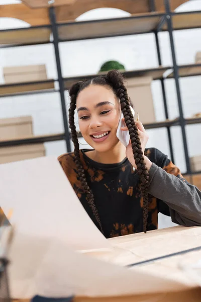 Smiling african american designer talking on smartphone and holding paper in workshop — Stock Photo