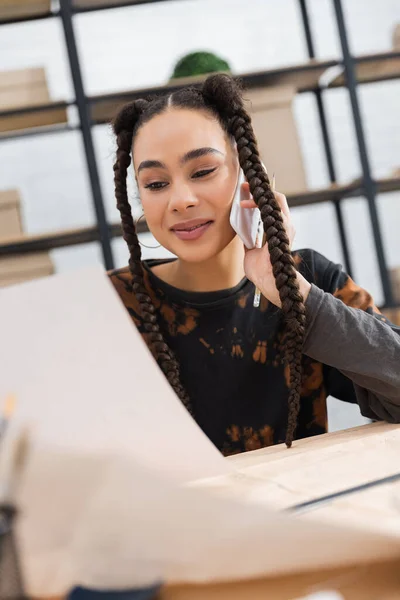 Улыбающаяся африканская американская ремесленница, глядя на размытую бумагу и разговаривая по телефону в мастерской — стоковое фото
