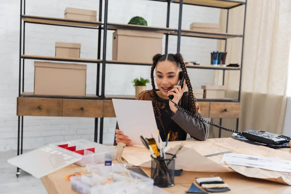 Smiling african american designer talking on telephone and holding sketch near sewing equipment — Stock Photo