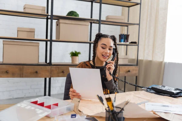 Smiling african american designer talking on telephone and holding sketch in workshop — Photo de stock