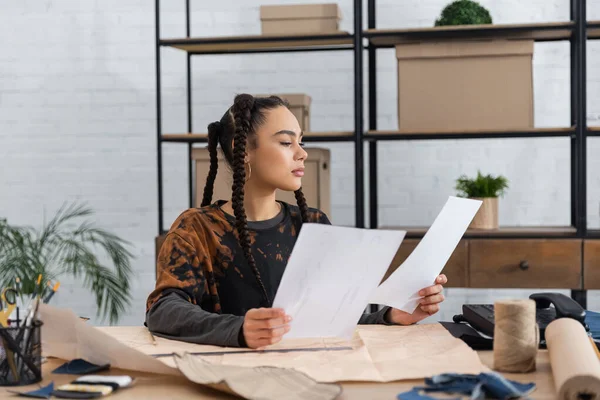 Focused african american designer holding papers near sewing equipment in workshop — Stock Photo