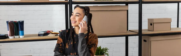 Cheerful african american designer talking on smartphone near carton boxes in workshop, banner - foto de stock