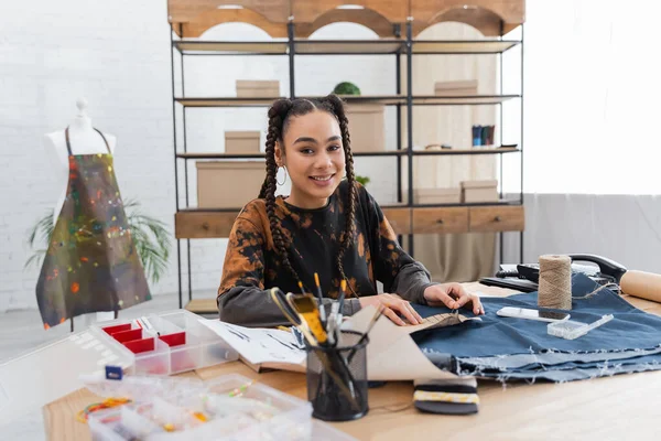 Smiling african american designer holding sewing pattern near cloth and decor on table in workshop — Foto stock