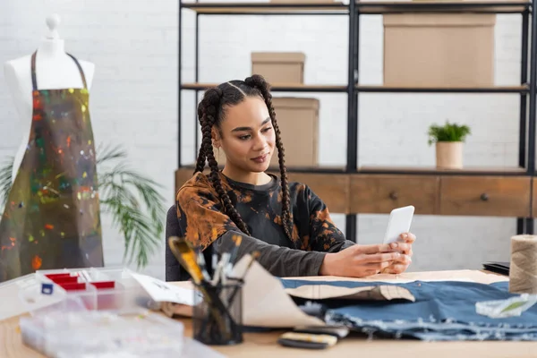 African american designer using smartphone near blurred sewing equipment in workshop — Stock Photo
