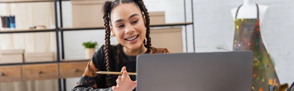 African american designer with pencil having video call on laptop in workshop, banner — Stock Photo