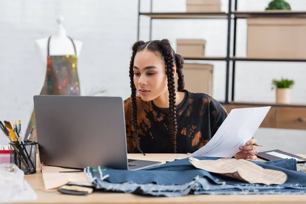 African american craftswoman holding sketch near laptop and cloth in workshop — Foto stock