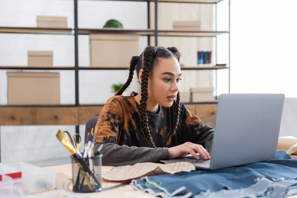 African american craftswoman using laptop near cloth in workshop — Stock Photo