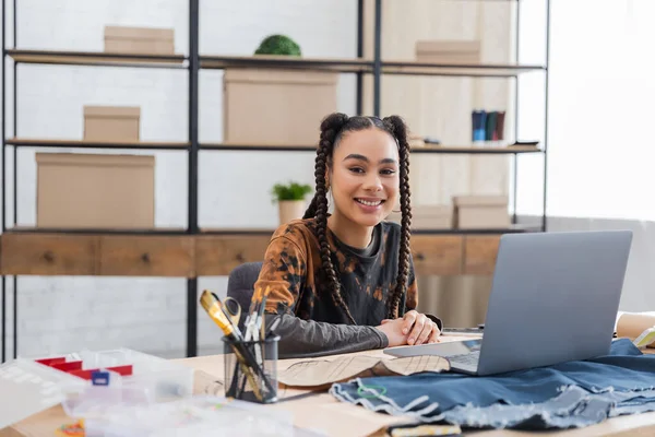 Smiling african american designer looking at camera near laptop and cloth in workshop — Foto stock