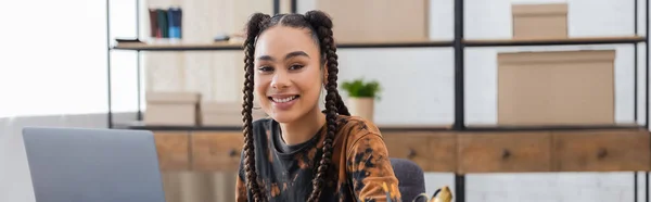 Cheerful african american designer looking at camera near laptop in workshop, banner — Foto stock