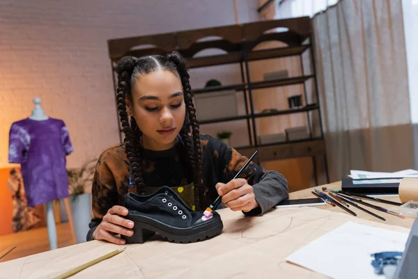 African american craftswoman painting on shoe near smartphone and sewing print in workshop — Fotografia de Stock