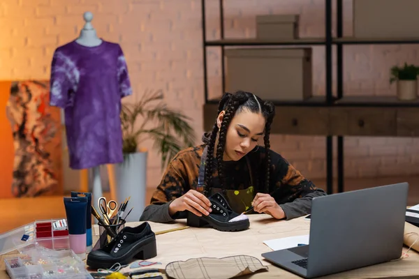 African american designer painting on shoe near laptop and sewing equipment — Stock Photo