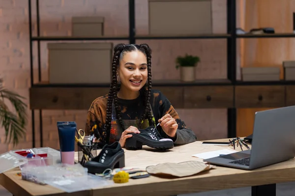Усміхнений афроамериканський дизайнер малює на взутті біля швейного обладнання — стокове фото