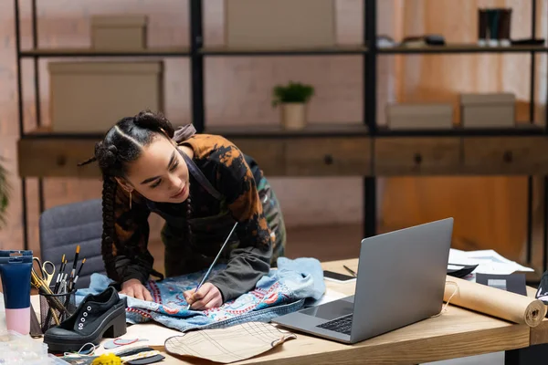 African american designer painting on denim jacket near laptop in workshop — Stock Photo