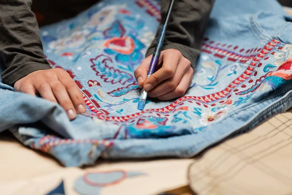 Cropped view of african american designer painting on denim jacket near blurred sewing pattern — Stock Photo