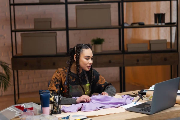 African american craftswoman holding cloth and looking at laptop in workshop — Foto stock