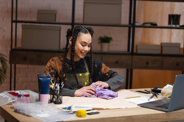 Весела афроамериканська майстриня тримає тканину біля швейного візерунка та пристроїв у майстерні — стокове фото