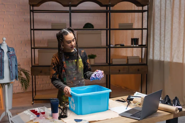 African american craftswoman holding cloth near box with recycle sign and devices in workshop — Fotografia de Stock