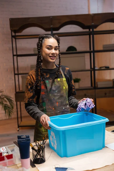 Smiling african american craftswoman holding cloth near box with recycle sign in workshop — Photo de stock