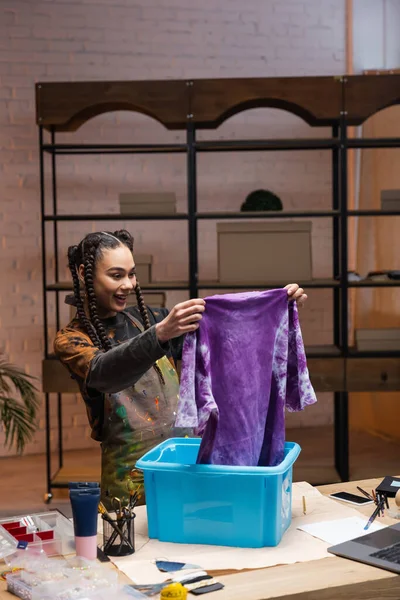 Smiling african americn designer holding t-shirt near container in workshop — Stock Photo