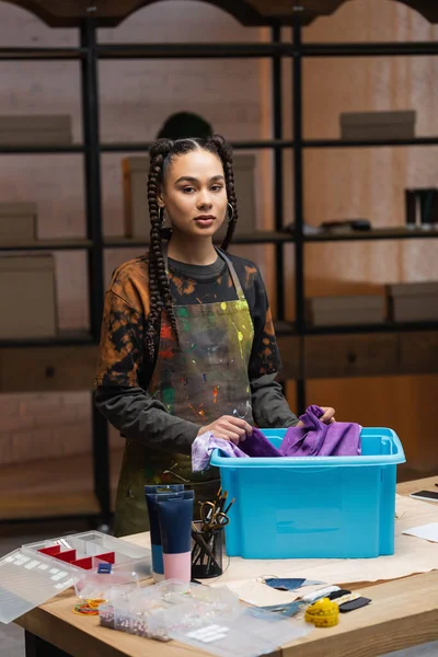 African american designer holding cloth near box and looking at camera in workshop — Stock Photo