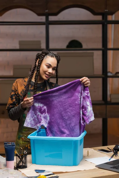 Cheerful african american craftswoman holding t-shirt in tie dye style near box in workshop — Photo de stock