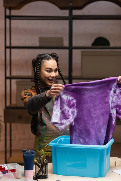 Positive african american craftswoman holding t-shirt in tie dye style near box in workshop — Stockfoto