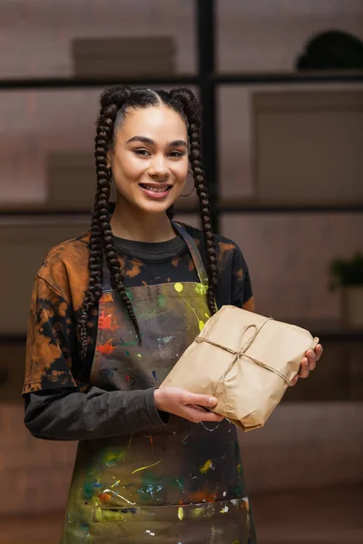 Positive african american designer in apron holding package and looking at camera in workshop — Foto stock