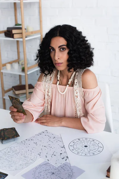 KYIV, UKRAINE - JUNE 29, 2022: brunette fortune teller holding tarot card and looking at camera near constellations charts — Stock Photo