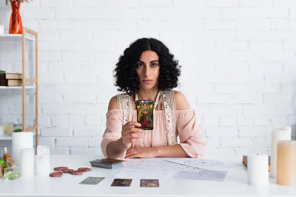 KYIV, UKRAINE - JUNE 29, 2022: fortune teller with tarot card looking at camera near star charts and clay runes — Fotografia de Stock