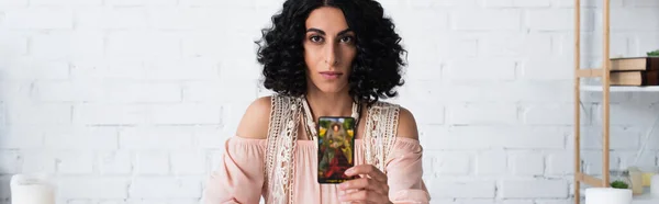 KYIV, UKRAINE - JUNE 29, 2022: brunette fortune teller holding tarot card and looking at camera at home, banner — Photo de stock