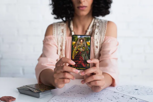 KYIV, UKRAINE - JUNE 29, 2022: cropped view of blurred predictor holding tarot card near celestial charts — стоковое фото