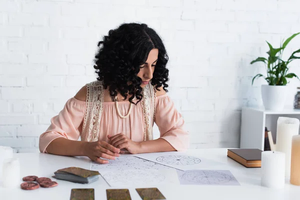 KYIV, UKRAINE - JUNE 29, 2022: brunette fortune teller looking at natal charts near tarot cards and clay runes — Foto stock