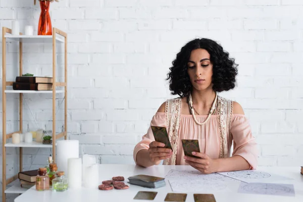 KYIV, UKRAINE - JUNE 29, 2022: young brunette woman predicting on tarot cards near clay runes and candles at home — Photo de stock