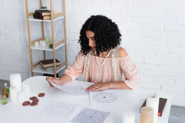 Astrologer looking at constellation charts near clay runes and candles — стоковое фото