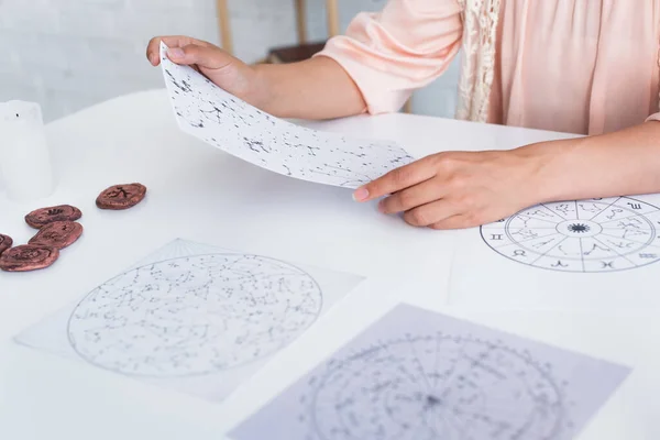 Cropped view of astrologer holding star charts near clay runes on table — Photo de stock