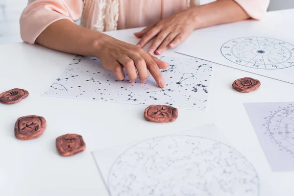 Cropped view of astrologer pointing at constellation charts near clay runes — стоковое фото